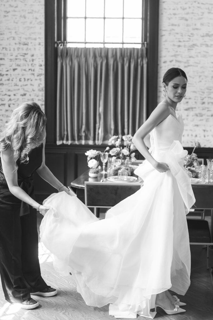 This black-and-white image captures a high-end luxury wedding planner attentively assisting a bride with her elegant gown. The scene is set in a beautifully decorated room featuring sophisticated decor elements, including a tastefully arranged dining table adorned with flowers and fine china. The bride, wearing a stunning, voluminous white wedding dress, looks serene and graceful as the planner ensures every detail of her attire is perfect. The planner's focus on the bride's train highlights her dedication to providing exceptional service and meticulous attention to detail. This image embodies the essence of luxury wedding planning, showcasing the planner's commitment to creating a flawless and memorable wedding experience.