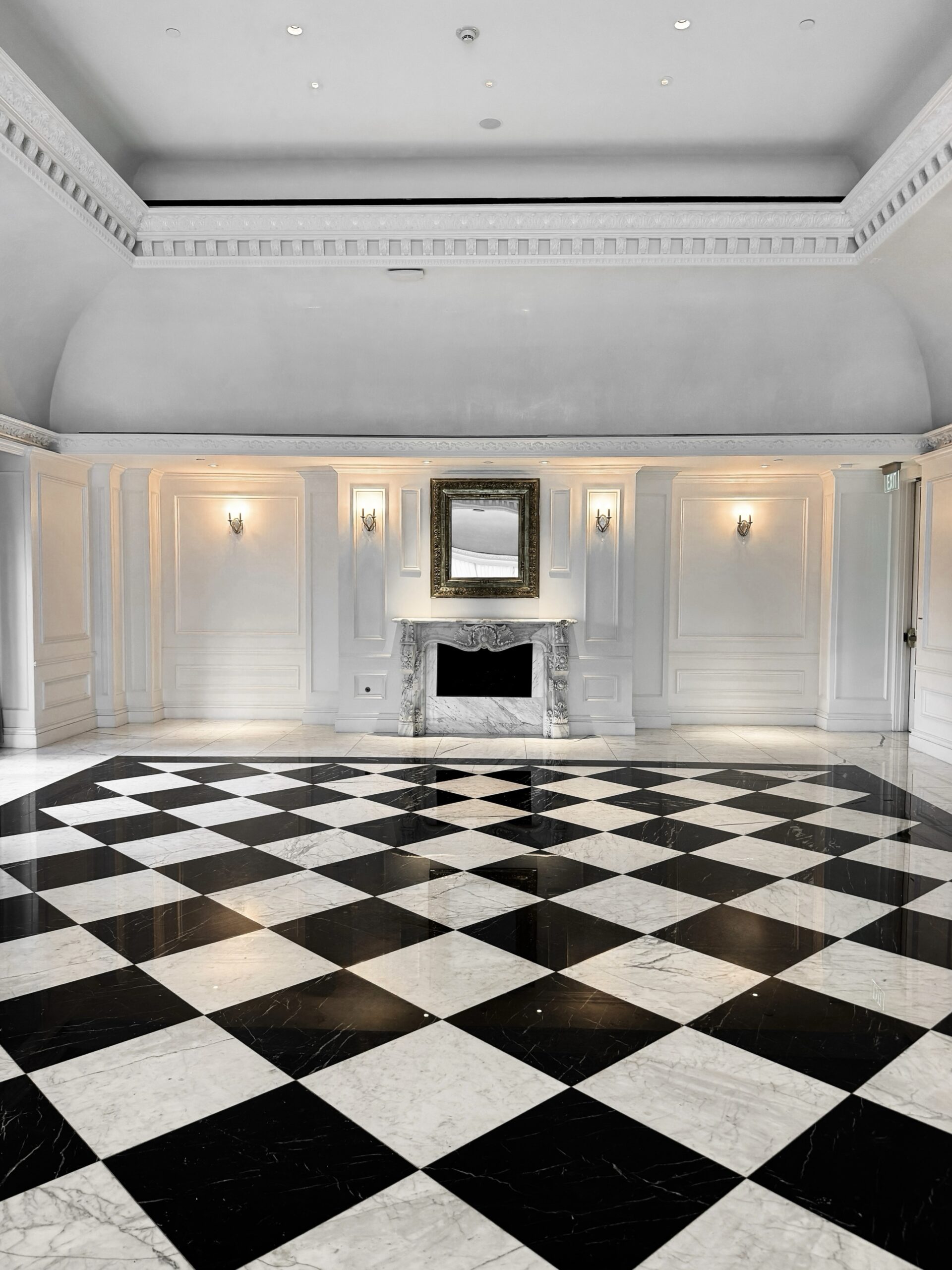 This image depicts an elegant and luxurious interior room with a high, vaulted ceiling and ornate moldings. The focal point is a grand, black-and-white checkered marble floor that leads to a decorative marble fireplace adorned with an ornate gold-framed mirror above it. Wall sconces provide soft, ambient lighting, highlighting the intricate details of the paneled walls. The overall aesthetic of the room is classical and sophisticated, reminiscent of a grand ballroom or a high-end hotel lobby. The polished marble floor reflects the lighting, adding to the opulent atmosphere.