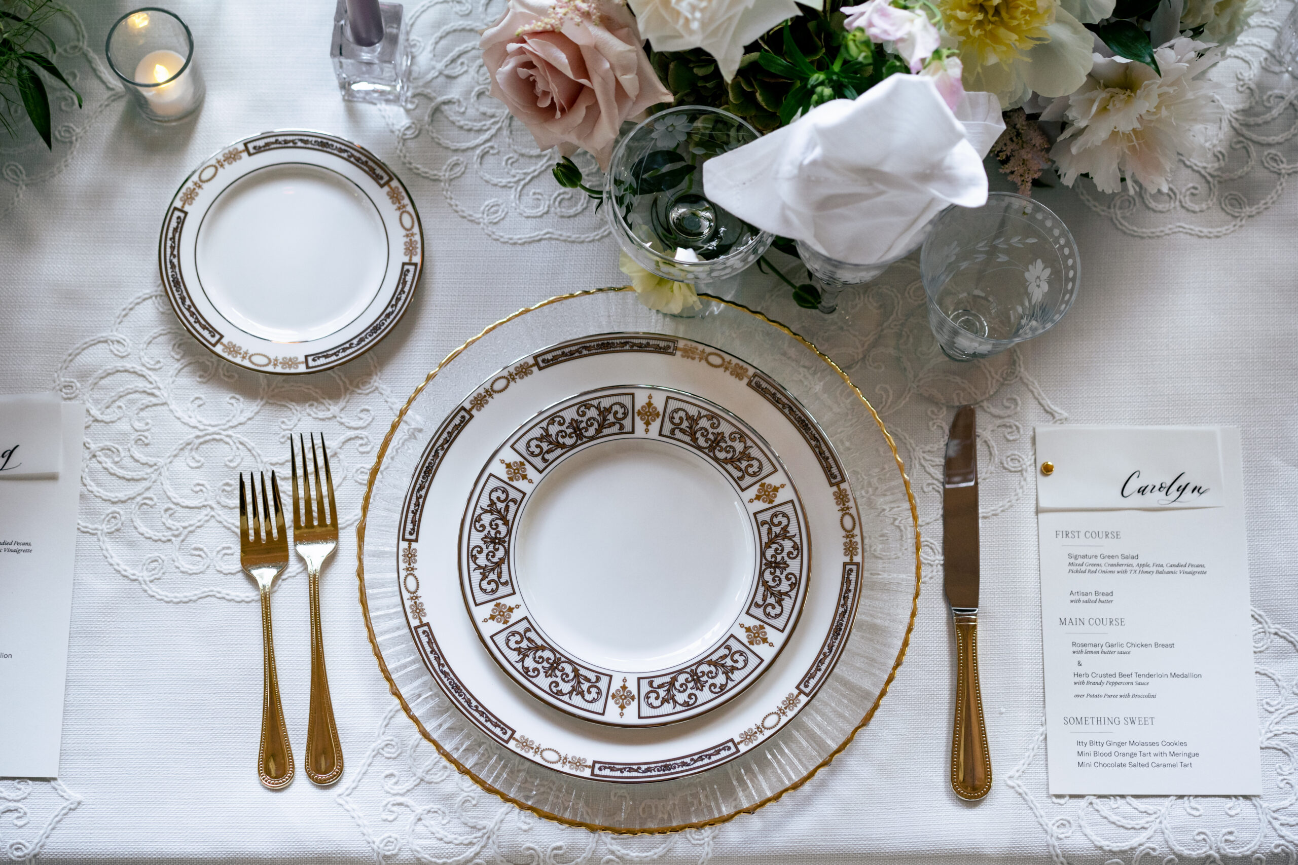 This image features an elegantly set dining table with a sophisticated place setting. The table is covered with a finely embroidered white tablecloth. The centerpiece of the setting is a decorative china plate with intricate gold and black patterns, accompanied by a smaller side plate with matching designs. Gold-plated cutlery, including two forks and a knife, is arranged neatly alongside the plates. A crystal glass with an etched floral design holds a white napkin, and fresh flowers in soft pastel colors add a touch of elegance. A menu card with the name "Carolyn" details the courses for the meal, including a first course, main course, and desserts. A lit candle in a small glass holder enhances the intimate and refined ambiance of the table setting.