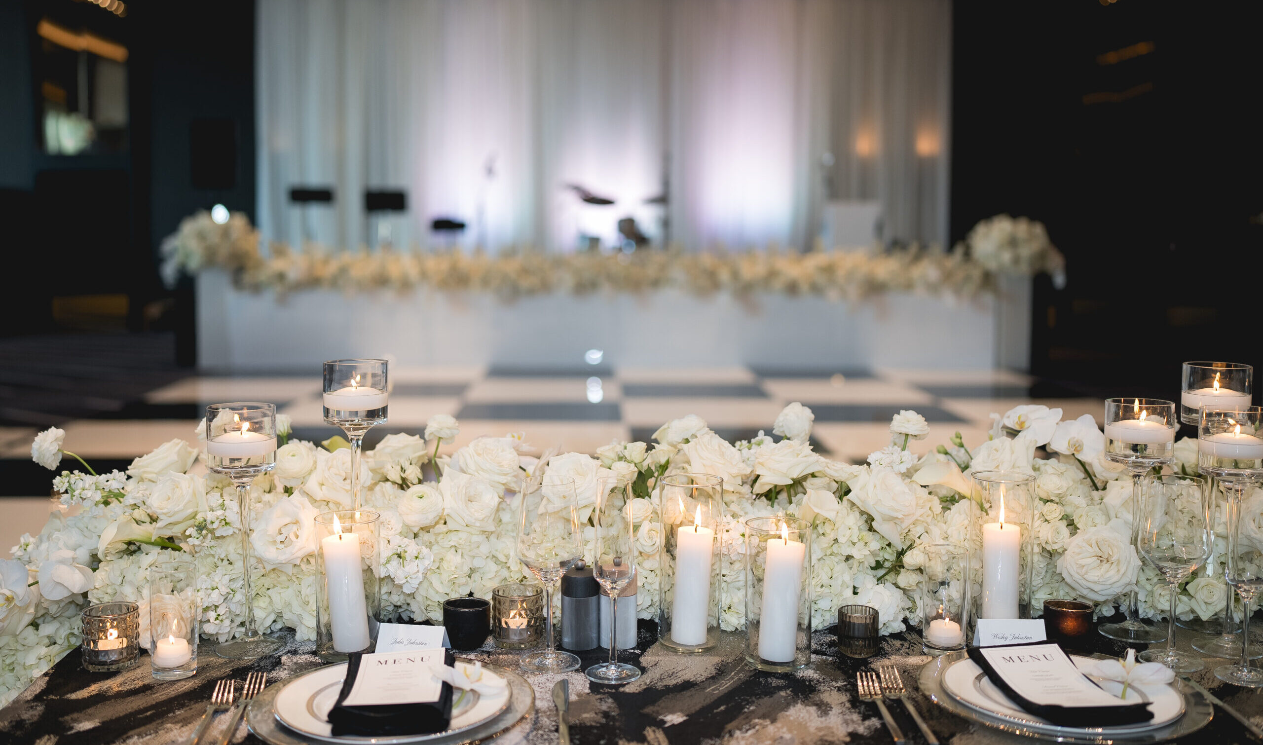 Stunning reception table setup at a Dallas luxury wedding, featuring a dramatic white floral centerpiece with roses and orchids, glowing candlelight in glass holders, modern black and white table settings with custom menus, and a view of a chic draped stage with a checkerboard dance floor in the background.